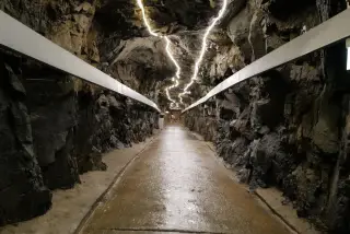 Ilustrasi: Lorong terowongan bawah tanah dengan dinding batuan cadas dan lampu penerangan memanjang.