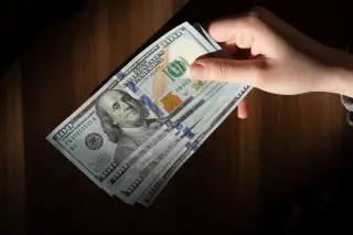 Ilustrasi: Tangan seseorang memegang beberapa lembar uang kertas seratus dolar di atas meja kayu.
