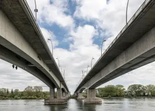 Ilustrasi: Dua jembatan beton kembar melintasi sungai berair tenang di bawah langit berawan.