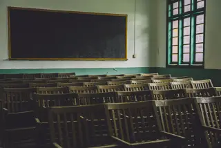 Ilustrasi: Deretan kursi kayu kosong di dalam ruang kelas dengan papan tulis hitam di dinding.