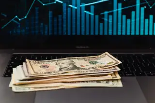 Ilustrasi: Tumpukan uang kertas dolar AS di atas papan ketik laptop dengan latar grafik keuangan.