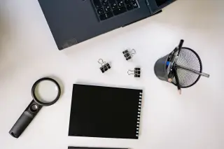 Ilustrasi: Tampilan atas meja kerja dengan laptop, buku catatan hitam, kaca pembesar, dan alat tulis.