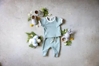 Ilustrasi: Setelan baju bayi hijau muda, sepatu kecil, dan mainan rajut tertata di atas lantai semen.