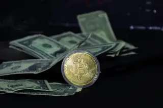 Ilustrasi: Koin emas Bitcoin bersandar di depan tumpukan uang kertas dolar AS dengan latar gelap.