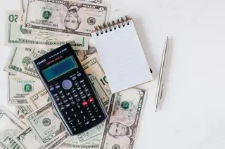 Ilustrasi: Kalkulator, buku catatan kosong, dan pulpen di atas tumpukan uang kertas dolar AS.