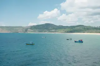 Ilustrasi: Beberapa perahu nelayan mengapung di laut biru dekat garis pantai berbukit hijau di bawah langit cerah.