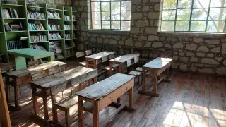 Ilustrasi: Ruang kelas kosong dengan meja dan bangku kayu serta rak buku di dekat dinding batu.