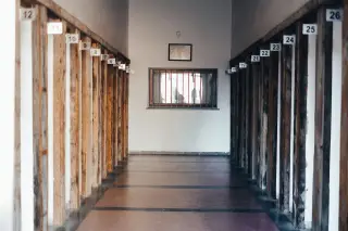 Ilustrasi: Lorong bangunan kosong dengan tiang-tiang kayu bernomor dan jendela berterali di ujungnya.