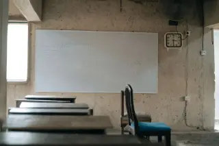 Ilustrasi: Ruang kelas kosong dengan deretan meja kayu dan papan tulis putih di dinding.