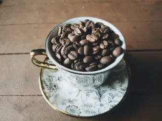 Ilustrasi: Secangkir keramik bermotif terisi penuh biji kopi sangrai di atas meja kayu.