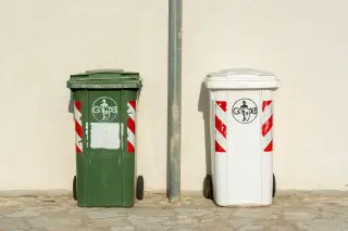 Ilustrasi: Dua tempat sampah beroda warna hijau dan putih bersandar di dinding luar ruangan.