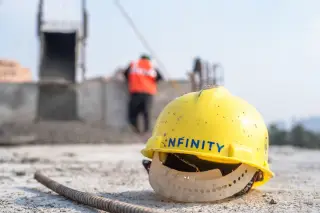 Ilustrasi: Sebuah helm proyek berwarna kuning tergeletak di area konstruksi dengan pekerja di latar belakang.