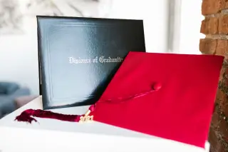 Ilustrasi: Topi toga merah dan map ijazah bertuliskan Diploma of Graduation di atas permukaan putih.