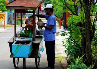 Ilustrasi: Seorang pedagang keliling berkaus biru menyiapkan makanan di samping gerobaknya di tepi jalan.