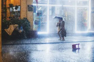 Ilustrasi: Dua orang berjalan di bawah payung di tengah hujan deras di depan pertokoan pada malam hari.