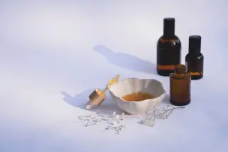 Ilustrasi: Botol kaca cokelat, pipet tetes, dan mangkuk keramik kecil di atas latar putih.