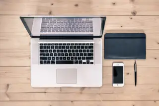 Ilustrasi: Laptop, ponsel pintar, buku catatan hitam, dan pena di atas meja kayu tampak atas.