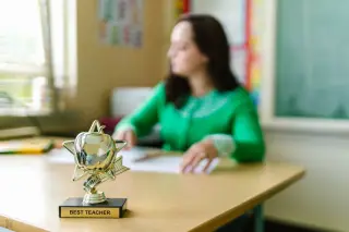 Ilustrasi: Piala bertuliskan Best Teacher di atas meja dengan sosok guru wanita di latar belakang buram.