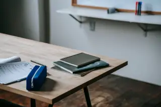 Ilustrasi: Meja belajar kayu dengan tumpukan buku catatan, lembar kerja, dan kotak pensil di dalam kelas.