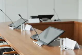 Ilustrasi: Meja rapat kayu dengan layar monitor, mikrofon konferensi, dan gelas kosong.