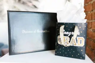 Ilustrasi: Map ijazah hitam bertuliskan Diploma of Graduation bersanding dengan kantong hadiah kelulusan.