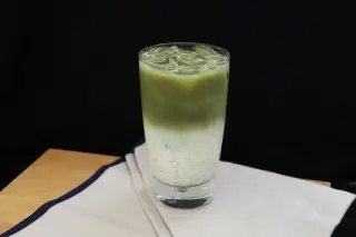 Ilustrasi: Segelas es matcha latte berlapis susu di atas serbet beralaskan meja kayu.