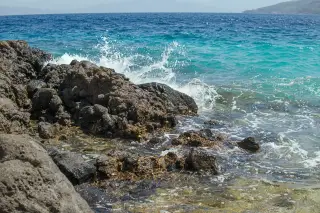 Ilustrasi: Ombak laut membentur bebatuan karang di tepi pantai dengan air laut biru.