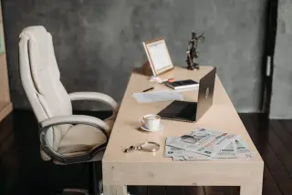 Ilustrasi: Meja kerja kayu dengan laptop terbuka, secangkir kopi, koran, dan kursi kantor kosong.