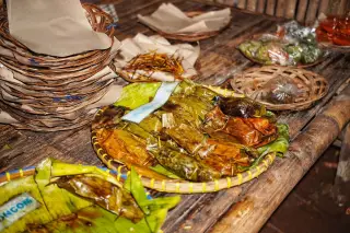 Ilustrasi: Makanan berbungkus daun pisang disajikan di atas wadah anyaman bambu di meja kayu.