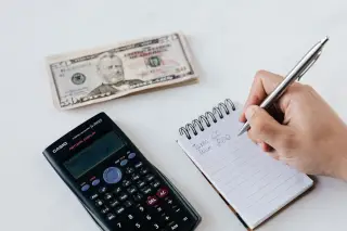 Ilustrasi: Tangan seseorang menulis di buku catatan di dekat kalkulator dan tumpukan uang kertas dolar AS.
