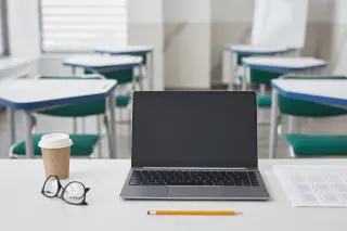 Ilustrasi: Laptop, kacamata, pensil, dan cangkir kopi di atas meja ruang kelas yang kosong.