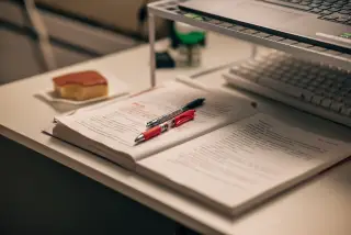 Ilustrasi: Buku catatan terbuka dengan dua pulpen di atas meja belajar dekat laptop.