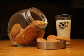 Ilustrasi: Toples kaca berisi biskuit dan segelas susu di atas meja kayu dengan latar gelap.