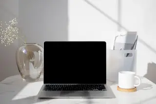 Ilustrasi: Laptop terbuka berlayar hitam di atas meja bersama cangkir dan vas bunga.
