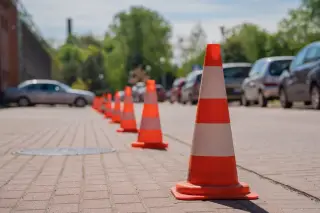 Ilustrasi: Deretan kerucut lalu lintas oranye bergaris putih di area jalan dekat kendaraan yang terparkir.