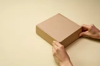 Ilustrasi: Sepasang tangan memegang kotak kardus cokelat polos di atas permukaan berwarna krem.
