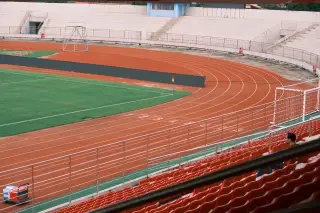 Ilustrasi: Lintasan lari atletik berwarna merah mengelilingi lapangan rumput di sebuah stadion kosong.
