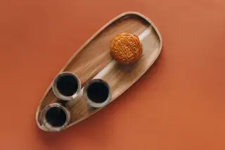 Ilustrasi: Sebuah kue bulan dan tiga cangkir kayu di atas nampan kayu berlatar polos cokelat.