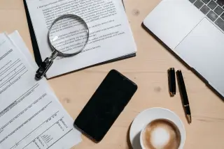 Ilustrasi: Dokumen, kaca pembesar, ponsel pintar, dan secangkir kopi di atas meja kerja.