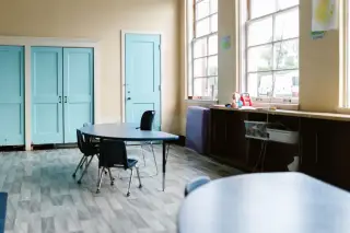 Ilustrasi: Ruang kelas TK atau sekolah dasar yang kosong dengan meja biru dan jendela besar.