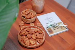 Ilustrasi: Kue kering dalam wadah anyaman bambu di meja kayu dekat buku dan kacamata.