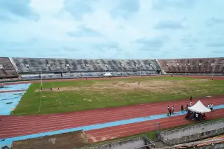 Ilustrasi: Stadion olahraga dengan lintasan lari merah dan lapangan rumput di bawah langit berawan.