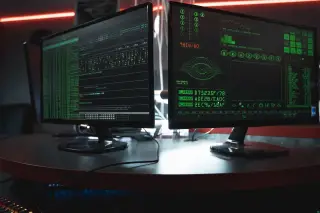 Ilustrasi: Dua layar monitor komputer menampilkan kode dan grafik hijau di ruangan bertabur cahaya neon.