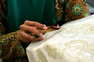 Ilustrasi: Tangan seseorang sedang memegang canting untuk menggambar motif pada kain batik.