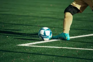 Ilustrasi: Kaki pesepak bola bersiap menendang bola di atas rumput lapangan hijau.