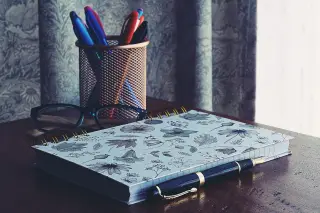 Ilustrasi: Buku catatan bermotif bunga, kacamata, pulpen, dan tempat pensil di atas meja kayu.
