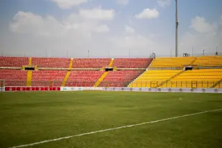 Ilustrasi: Pemandangan lapangan rumput dan deretan bangku tribun stadion olahraga yang kosong di siang hari.