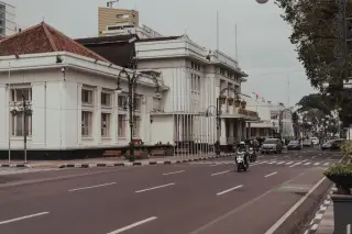 Ilustrasi: Pemandangan jalan raya beraspal dengan bangunan bergaya kolonial dan kendaraan yang melintas.