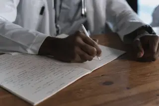 Ilustrasi: Seseorang berjas dokter menulis catatan di atas buku tulis beralaskan meja kayu.