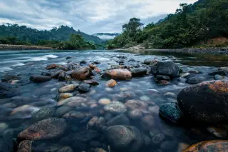 Ilustrasi: Aliran sungai berbatu dengan latar belakang perbukitan hijau yang berkabut.